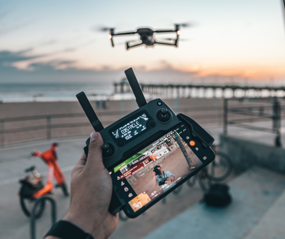 Dronen kauko-ohjain. Taustalla näkyy drone lennossa rantaa kohden.